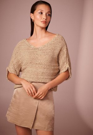Beige knitted top with a V-neck and rolled sleeves, paired with a light beige wrap skirt featuring a subtle sheen and smooth texture.