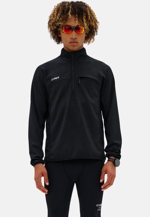 Persona che indossa un top sportivo nero a maniche lunghe e leggings, occhiali sportivi con lenti tinte di rosso e un orologio da polso nero, in piedi davanti a uno sfondo bianco.