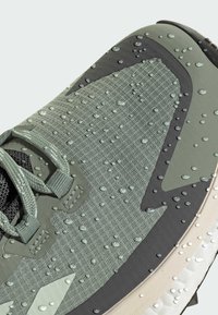 Waterdichte groene textiele sportschoen met een gestructureerd oppervlak, zwarte accenten en zichtbare waterdruppels op het bovenmateriaal.