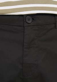 Černé kalhoty s předním zapínáním na knoflík a hladkým povrchem. Pas je částečně zakrytý pruhovaným béžovým a bílým topem.