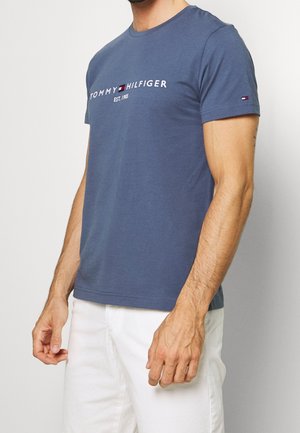 Mann trägt ein blaues Tommy Hilfiger T-Shirt mit Logo und weiße Hose vor einem einfarbigen hellen Hintergrund.
