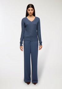 Niebieski sweter w serek z teksturowanym splotem, połączony z spodniami o wysokim stanem i szerokimi nogawkami w dopasowanym niebieskim kolorze oraz brązowymi spiczastymi butami.