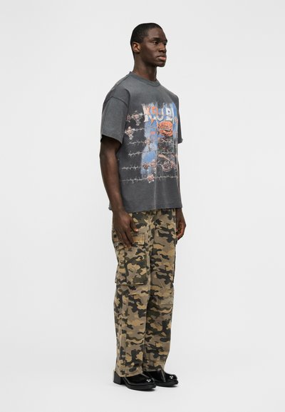 Γκρι γραφικό T-shirt με άνετη εφαρμογή, με σχέδιο λουλουδιών και συρματοπλέγματος, συνδυασμένο με μπεζ παραλλαγής παντελόνι cargo και μαύρα παπούτσια.