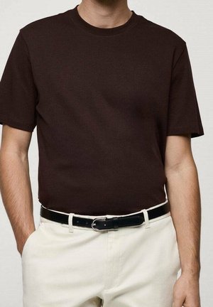 Persoon draagt een donkerbruin shirt met korte mouwen in een off-white broek met een zwarte riem, handen gedeeltelijk in de zakken.