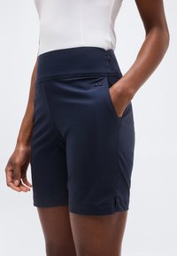 Γυναίκα που φοράει μπλε σκούρα σορτς υψηλής μέσης με πλαϊνές τσέπες και λευκό αμάνικο τοπ, όρθια με το ένα χέρι στην τσέπη.