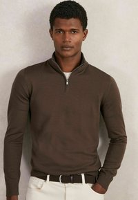 Brauner Reißverschluss-Pullover mit langen Ärmeln, glattem Strickstoff und einem dezenten Schimmer. Kombiniert mit cremefarbenen Hosen und einem gewebten braunen Gürtel.