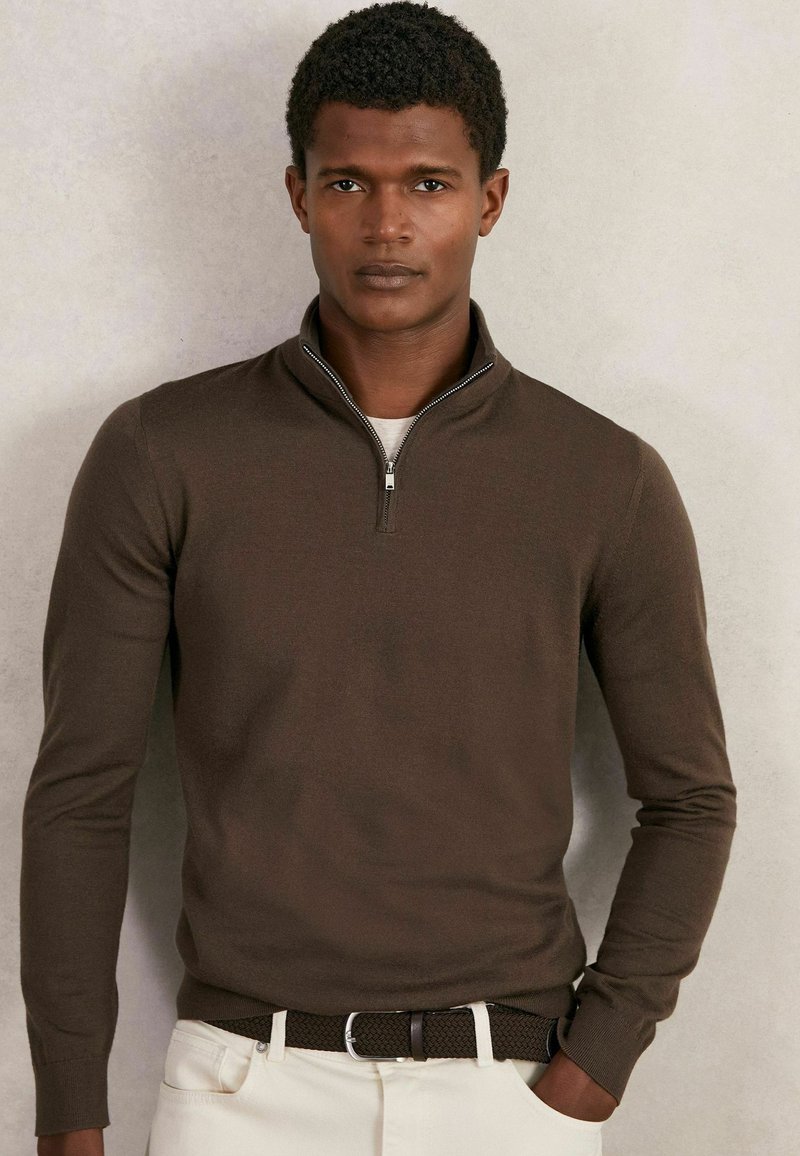 Brauner Reißverschluss-Pullover mit langen Ärmeln, glattem Strickstoff und einem dezenten Schimmer. Kombiniert mit cremefarbenen Hosen und einem gewebten braunen Gürtel.