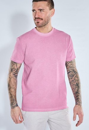 Mann mit Ärmeltattoos trägt ein schlichtes rosa kurzärmeliges T-Shirt und helle Hosen, blickt nach links.