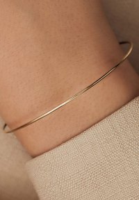 Dunne gouden metalen bangle rustend op een pols, met een gladde oppervlakte en eenvoudig ontwerp, in contrast met een gestructureerde beige stof.