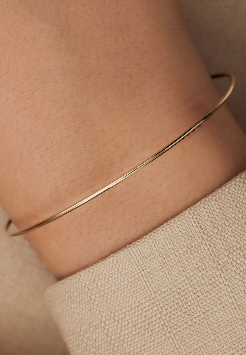 Dunne gouden metalen bangle rustend op een pols, met een gladde oppervlakte en eenvoudig ontwerp, in contrast met een gestructureerde beige stof.