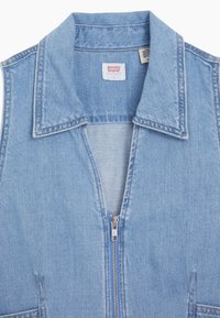 Colete de ganga azul claro com colarinho pontudo, fecho frontal em zíper e costura mínima. Apresenta dois bolsos laterais. Textura suave.