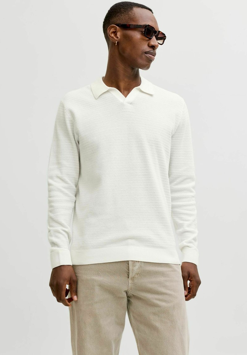 Pull-over blanc en tricot avec un col en V et un motif texturé, associé à un pantalon beige. Le mannequin porte des lunettes de soleil foncées.