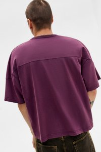 Man met kort haar, gekleed in een losse paarse shirt met korte mouwen en donkere broek, staand met de rug naar de camera tegen een effen lichte achtergrond.
