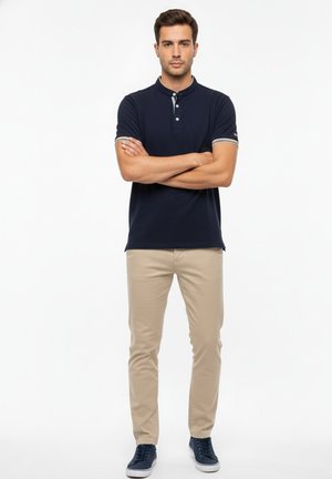 Hombre de pie con los brazos cruzados, vestido con un polo de manga corta azul marino, pantalones beige y zapatillas azul marino, sobre un fondo blanco liso.