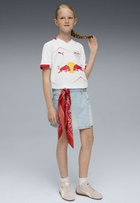 Camiseta blanca de manga corta con detalles en rojo, con un logo de toro rojo y un círculo amarillo, combinada con una falda de mezclilla clara y un pañuelo rojo.