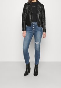 Persona con chaqueta de cuero negra de motociclismo, camiseta negra, vaqueros ajustados de tiro alto en blue desgastados con cierre de botones, y botines negros.