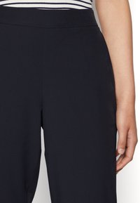 Námořnické kalhoty s hladkou texturou, s vysokým pasem a volným střihem. Ruka odpočívající poblíž bočního švu pro kontext.