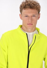 Chaqueta de cremallera amarilla neón con cuello alto, confeccionada en una tela suave. Presenta una cremallera negra y un detalle de escote en capas.