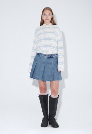 Junge Frau trägt einen weiß-blau gestreiften Pullover, blauen plissierten Jeansrock und schwarze kniehohe Stiefel und steht vor einem schlichten weißen Hintergrund.