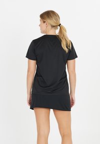 Nainen, jolla on vaaleat poninhäntä ja musta lyhythihainen paita sekä musta hame, seisoo valkoista taustaa vasten selin.