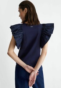 Blusa blu navy con maniche corte a ruffle. Tessuto morbido con una leggera lucentezza. La vista posteriore mostra il marchio sul colletto e le braccia incrociate.