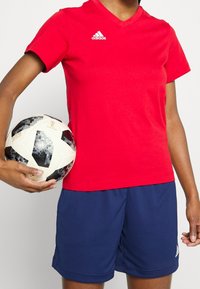 Röd kortärmad t-shirt med v-hals och vitt logotyp, tillsammans med blåa sportshorts; håller en mönstrad svart och vit fotboll.