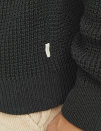 Sötét szürke, kötött pulóver, texturált szövettel, széles bordás mandzsettákkal és egy kis szövetcímkével az oldalon. Az alján kordbársony nadrág látható.