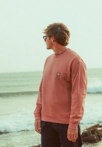 Sweat-shirt rose clair avec poignets et ourlet côtelés, arborant un petit motif floral sur la poitrine, porté avec un pantalon foncé par une personne à la plage.