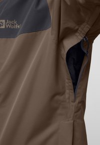 Bruine waterdichte jas met donkergrijze accenten, voorzien van een ritszak aan de zijkant en een gladde, matte textuur. Merken zichtbaar op de borst.