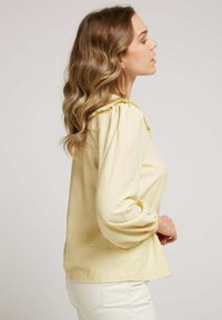 Femme de profil avec des cheveux châtain clair ondulés, portant un blouse à manches longues jaune pâle et un pantalon blanc, sur un fond uni.