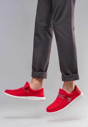 Person trägt hochgekrempelte, dunkelgraue Hosen und leuchtend rote Freizeitschuhe mit weißen Sohlen, in der Luft vor einem grauen Hintergrund.