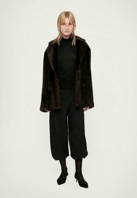 Bruine faux fur jas met een losse pasvorm, zachte textuur en brede revers. Gecombineerd met een zwarte coltrui en losse broek, wat de look compleet maakt.