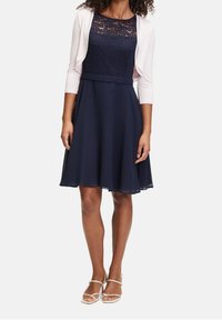 Vestido azul marino con cuerpo de encaje y falda acampanada, acompañado de un cárdigan blanco. El vestido presenta una cintura ajustada y una tela suave y fluida.