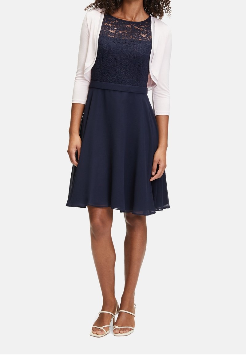 Vestido azul marino con cuerpo de encaje y falda acampanada, acompañado de un cárdigan blanco. El vestido presenta una cintura ajustada y una tela suave y fluida.