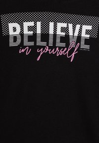 Camiseta negra con el texto "CREE en ti mismo" en blanco y rosa, con diferentes fuentes y patrones arriba. Textura de tela suave.