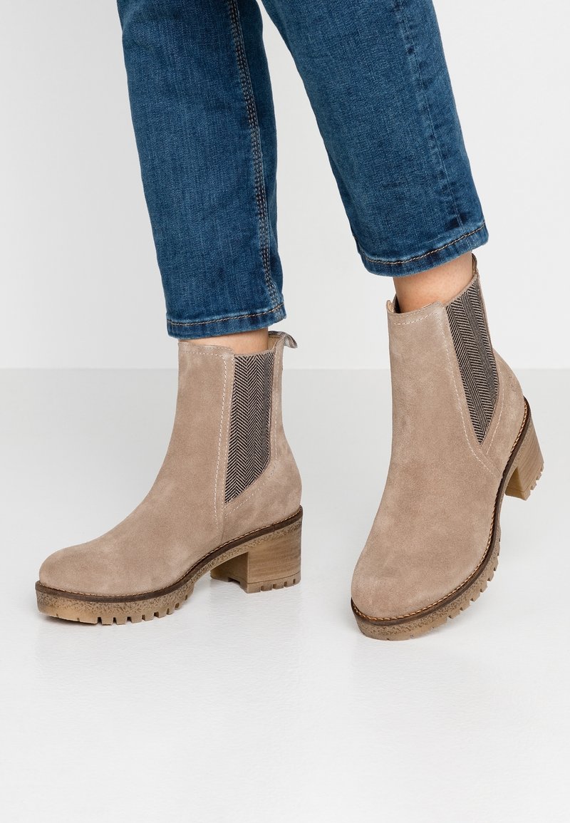 Pieds chaussés de bottines en daim brun clair à talons épais et panneaux latéraux élastiques, associées à un jean bleu raccourci.