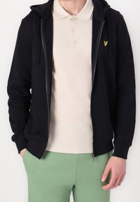 Černá zipová mikina s přední kapsou, pod ní béžová polokošile a zelené kalhoty. Tkanina vypadá hladce a má jemný detail s logem.