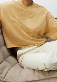 Persona vestida con un sudadera oversize beige que dice "MALIBU CALIFORNIA" y pantalones cargo blanco roto, sentada en un sofá de textura beige.