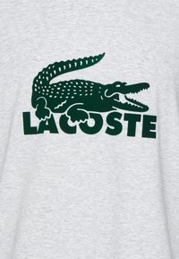 Grå T-shirt med en grön alligatorlogotyp och ordet "LACOSTE" i fet svart text. Mjukt material, avslappnad design.