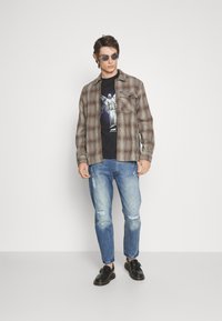 Rutig flanellskjorta i bruna nyanser, svart grafisk t-shirt, slitna blåjeans och svarta slip-on skor. Rundade solglasögon fulländar looken.