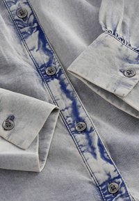 Denim shirt met een lichtblauw en grijs tie-dye patroon, voorzien van knoopsluiting aan de voorkant en mouwen, en donkergrijze knopen met bloemenontwerpen.