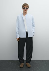Camicia button-up azzurro chiaro, t-shirt bianca sotto, pantaloni neri a gamba larga, scarpe slip-on marroni e occhiali da sole neri. Design semplice e moderno.