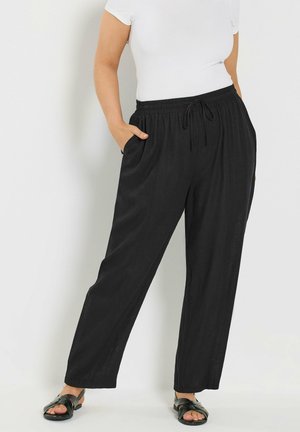 Frau trägt lockere schwarze Kordelzughose, weißes kurzärmeliges Oberteil und schwarze Sandalen, steht mit einer Hand in der Tasche.