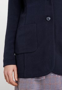 Námořnický modrý pletený cardigan s jedním knoflíkovým zapínáním, s velkou nášivkou na levé straně a zdobenými manžetami.