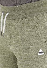 Pantaloni della tuta verde oliva con una finitura testurizzata, dotati di un laccio bianco e di una patch a forma di triangolo sul lato.