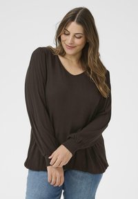 Kvinde med langt brunt hår iført en løs, mørkebrun bluse med lange ærmer og blå jeans, smilende blidt med lukkede øjne og hænderne foldet.