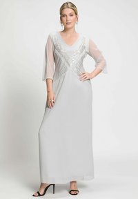 Maxi dress grigio chiaro con scollatura a V e maniche a campana trasparenti. Presenta ricami floreali argentati e pieghe verticali. Indossato con tacchi alti neri.