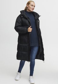Melna gara pufīga jaka ar kapuci, ar rāvējslēdzēja aizdari, sānu kabatām un quilted dizainu. Uzvilkta virs tumši zila topa un zila džinsu.