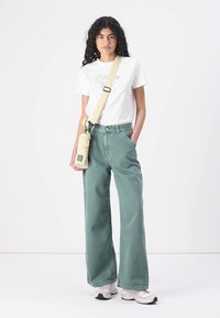 Lysegrønne vidde jeans laget av bomull, kombinert med en hvit bomullst-skjorte med trykt tekst og en beige crossbody-veske med justerbar stropp.