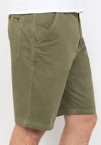 Pantalones cortos verde oliva hechos de algodón suave, con presillas para cinturón, bolsillos laterales y un bolsillo trasero de parche. Presenta una etiqueta con el logo en la cinturilla.